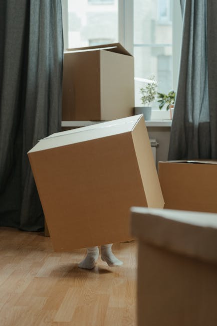 Inside a room with wooden flooring and large windows covered by dark grey curtains, several cardboard moving boxes are scattered, with one large box partially lifted by a person's legs visible beneath