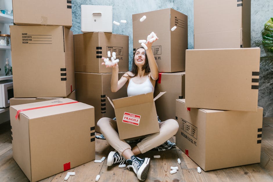 Inside a room with wooden flooring and large windows covered by dark grey curtains, several cardboard moving boxes are scattered, with one large box partially lifted by a person's legs visible beneath
